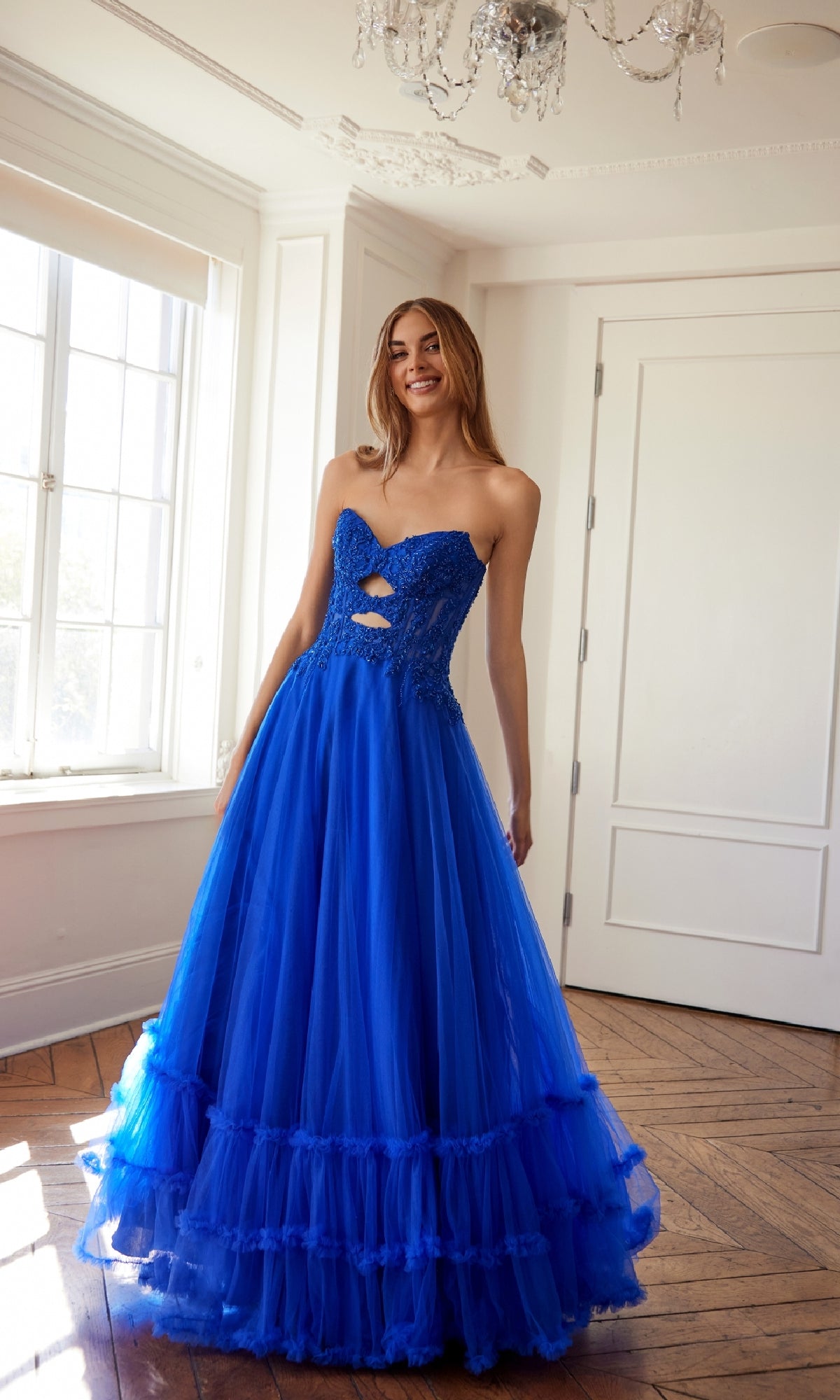 A woman stands indoors by a sunlit window, smiling in the Strapless Long Prom Ball Gown: Juliet JT2561S, featuring a floor-length royal blue tulle skirt, cutout details, and an embroidered lace bodice.